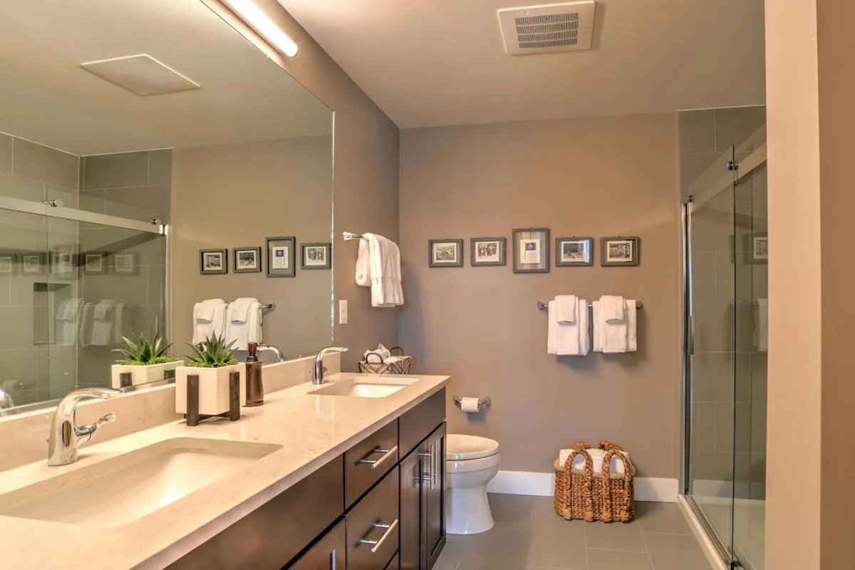 This image features a modern full bathroom, complete with a double sink vanity, framed photographs, and a shower stall. Morning sunlight reflects off the mirror, creating a calm and organized environment perfect for self-care routines.