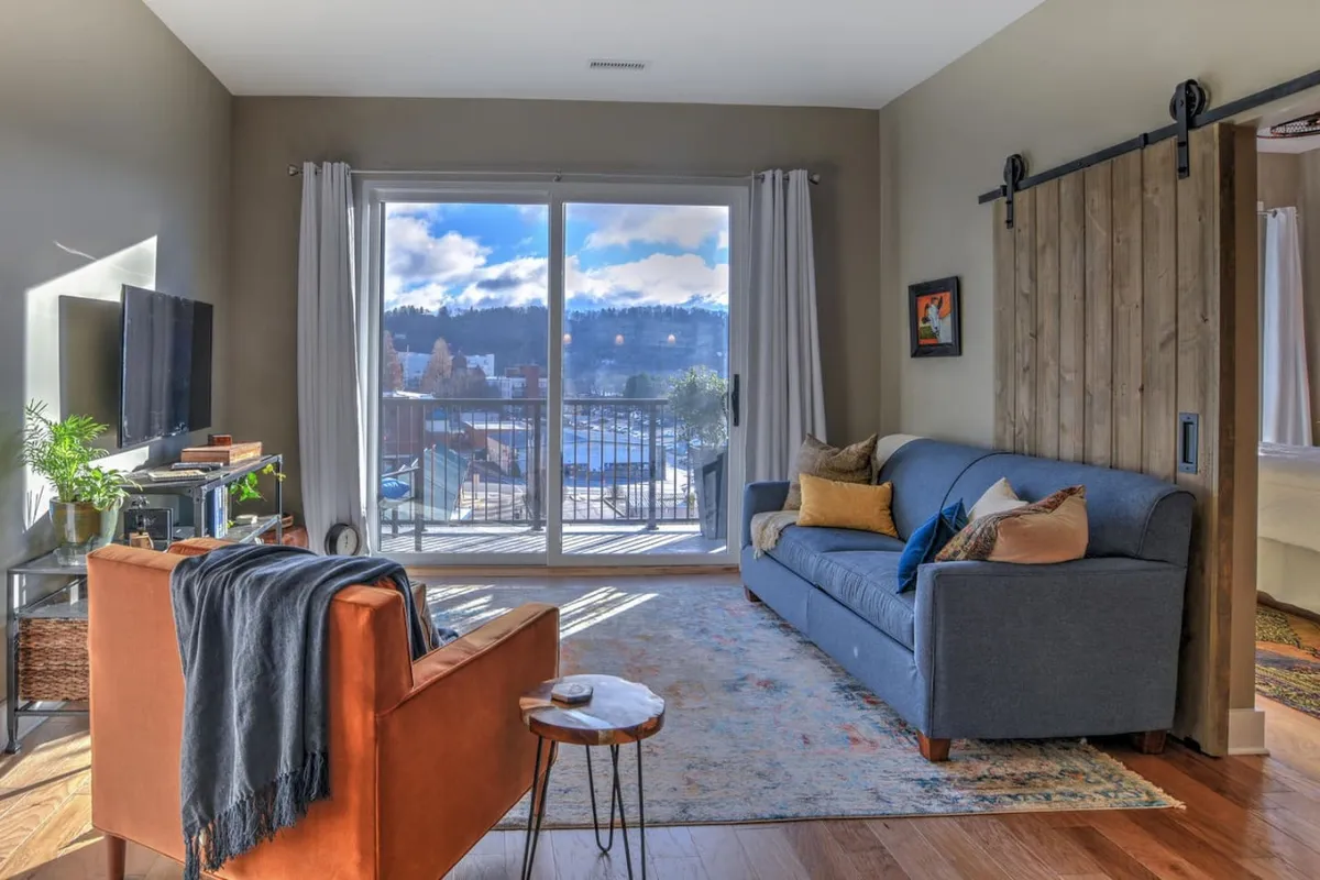A cozy and stylish open-concept living room featuring a comfortable sofa with colorful cushions, a wooden accent wall, and expansive windows that frame inspiring city views. The layout is ideal for gathering, relaxing, or hosting friends and family, with soft lighting and inviting decor.