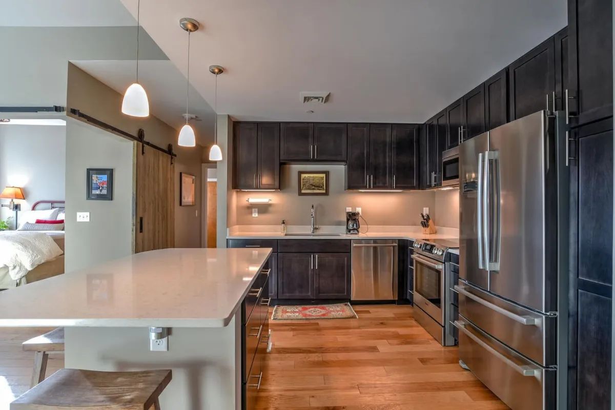 A spacious, modern kitchen featuring a central island with bar stools, gleaming stainless steel appliances, and warm wooden cabinetry. Pendant lighting casts a cozy glow, and the space is clearly designed for both cooking and gathering, with room for family or friends to connect.