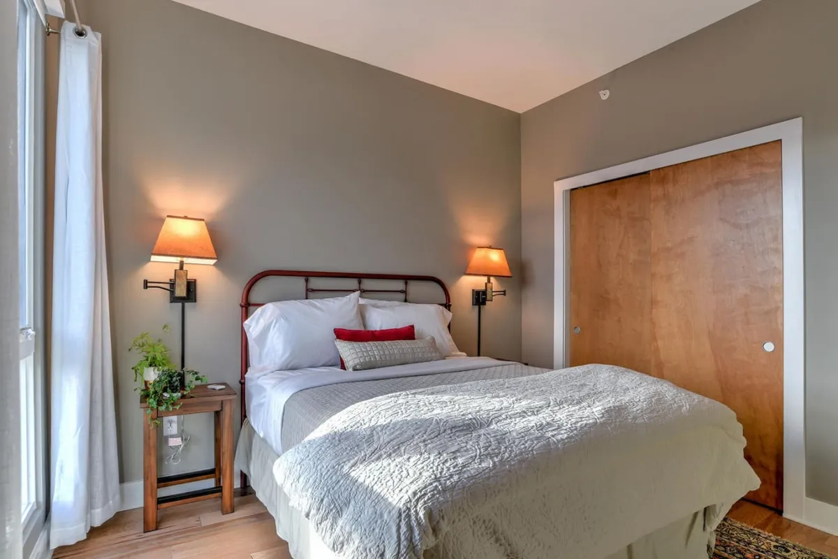 This image showcases a serene bedroom featuring a bed with decorative pillows, nightstands with lamps, a plant on the nightstand, and a wooden closet door. The space is bathed in soft morning light, creating a calm and restful atmosphere ideally suited for relaxation and rejuvenation.