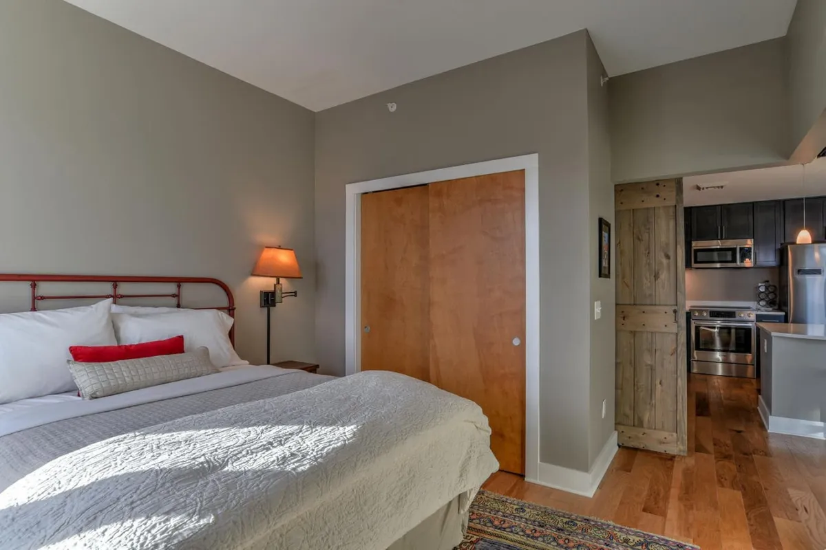 A serene bedroom featuring a plush bed, calming wall art, and the soft glow of morning sunlight. The view includes the entrance to the kitchen, creating a natural sense of flow and connectivity within the space—ideal for restful nights after exploring the city.