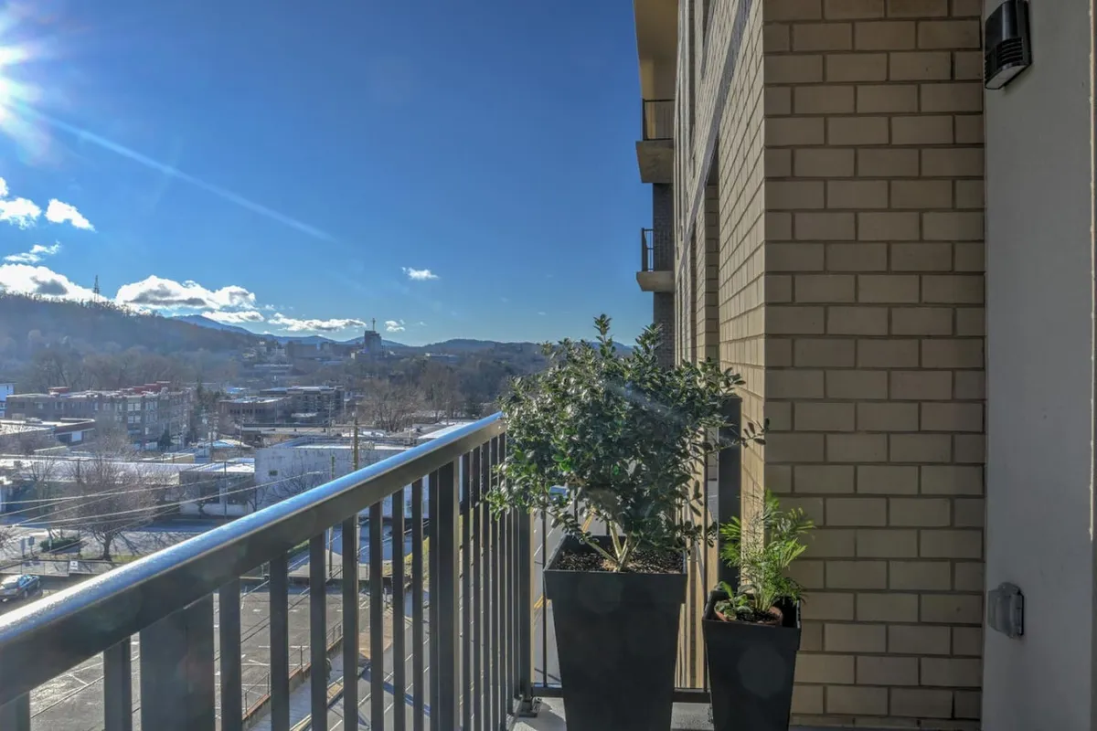 This image captures a private balcony adorned with lush potted plants and an expansive view of Asheville’s mountains beneath a broad sky. The serene outdoor space suggests a perfect spot for morning coffee, reading, or simply soaking in the fresh air while surrounded by the sights of the city and nature beyond.