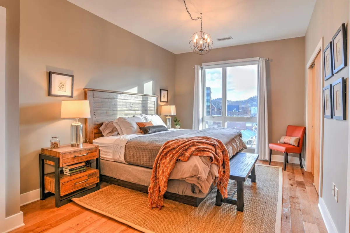 This image portrays a bedroom with textured bedding, a large window offering an inviting view, wooden nightstands, and a distinctive light fixture. The mood is serene and comfortable, suggesting a restorative home base for guests.