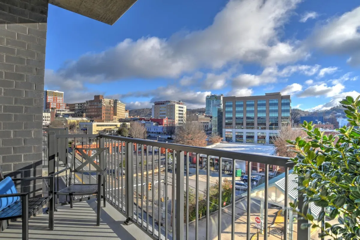 A balcony overlooking Asheville's city skyline and distant mountains, framed by a decorative railing and potted plants. The scene suggests a perfect spot for morning coffee, socializing, or soaking in the urban energy and natural beauty.