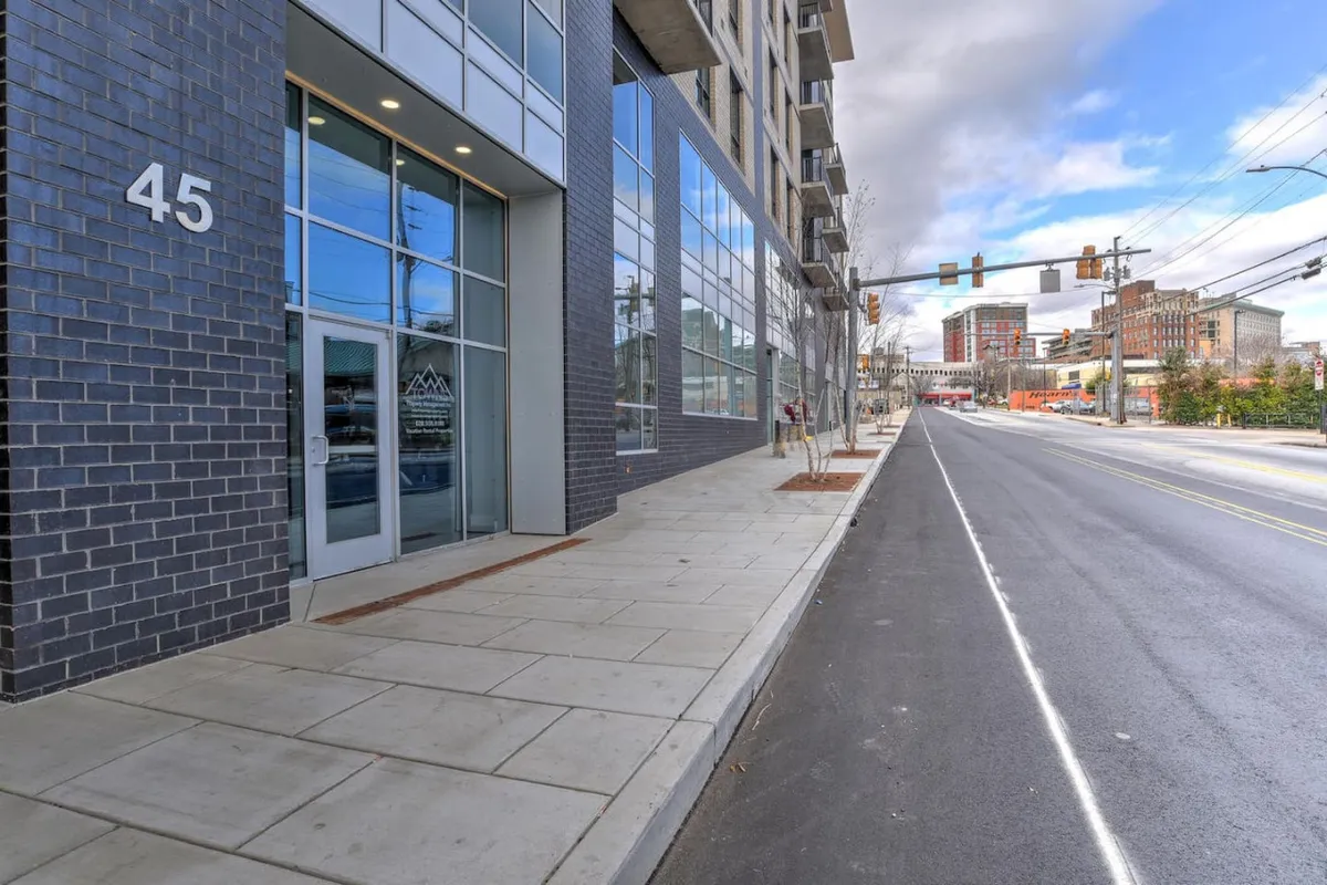 This image features the building's entrance with large glass doors and prominent building number, framed by expansive glass windows. The sidewalk is visible, suggesting easy access and an urban vibe.