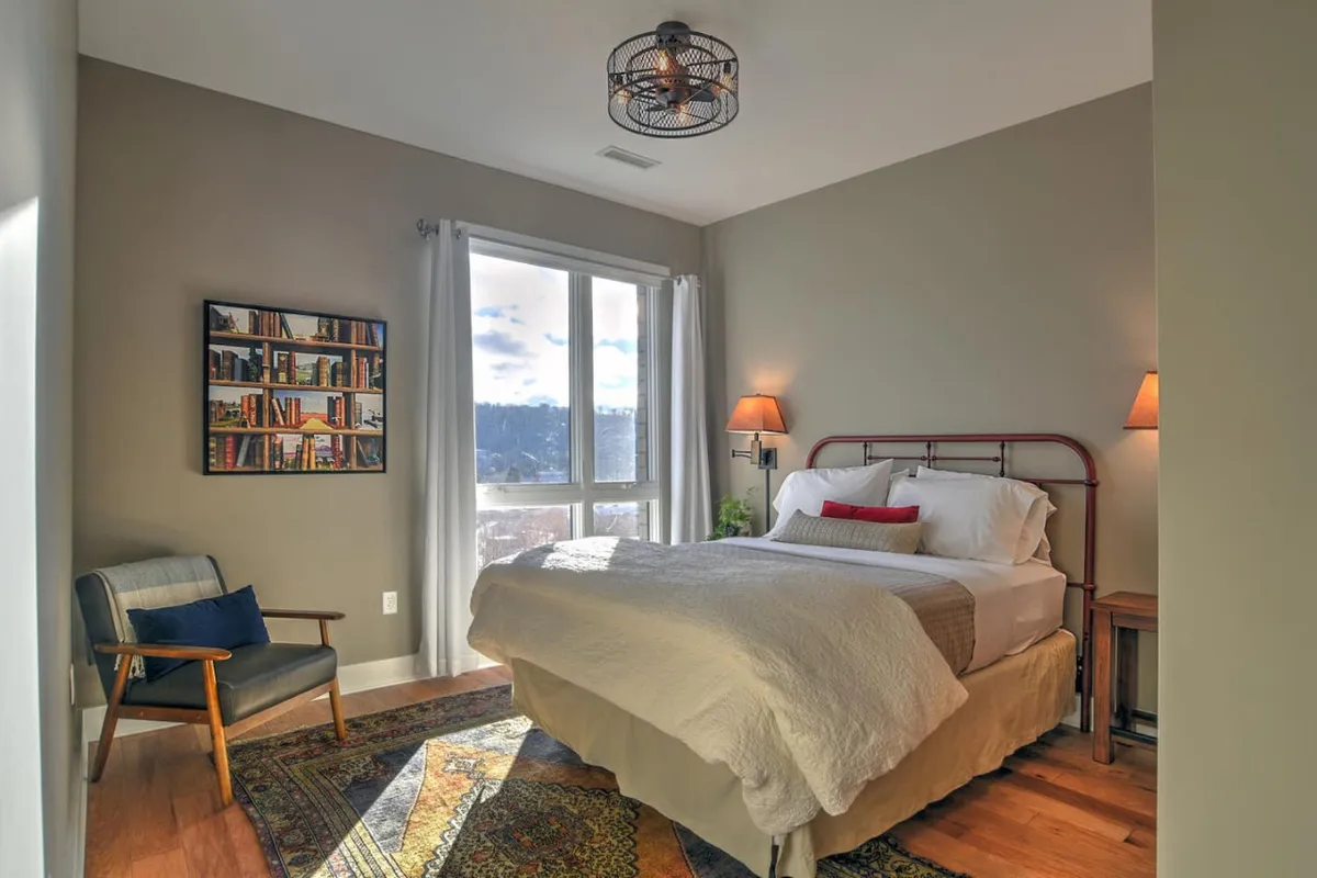 A cozy bedroom featuring a comfortable bed near a window that lets in natural light. The window provides a view outward, subtly suggesting the connection between indoor relaxation and the possibilities of exploring the city with your pet. A bookshelf adds a homey touch, making the space feel inviting for both guests and their four-legged companions.
